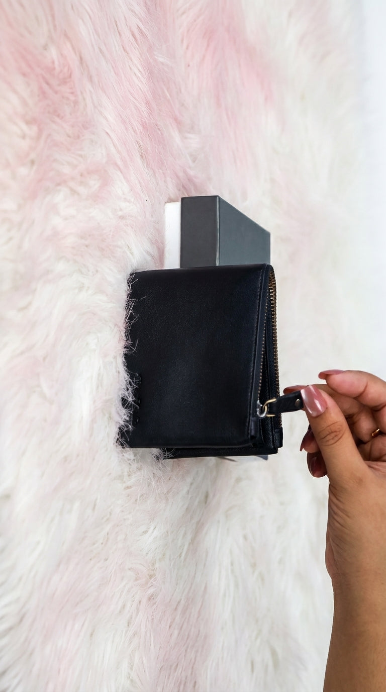 A black leather zippered wallet or card holder with a silver geometric accent, shown being held by a hand against a fluffy white and pink background.