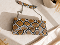 A snakeskin-patterned clutch bag with a matching wristlet strap and silver chain, styled on a light beige stone surface with soft shadows and dried grass accents.