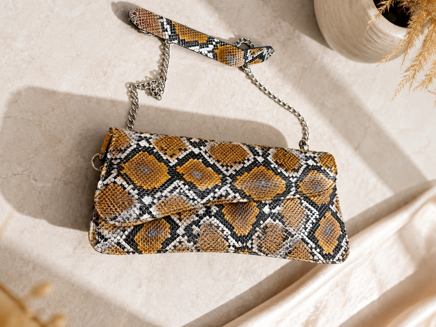 A snakeskin-patterned clutch bag with a matching wristlet strap and silver chain, styled on a light beige stone surface with soft shadows and dried grass accents.