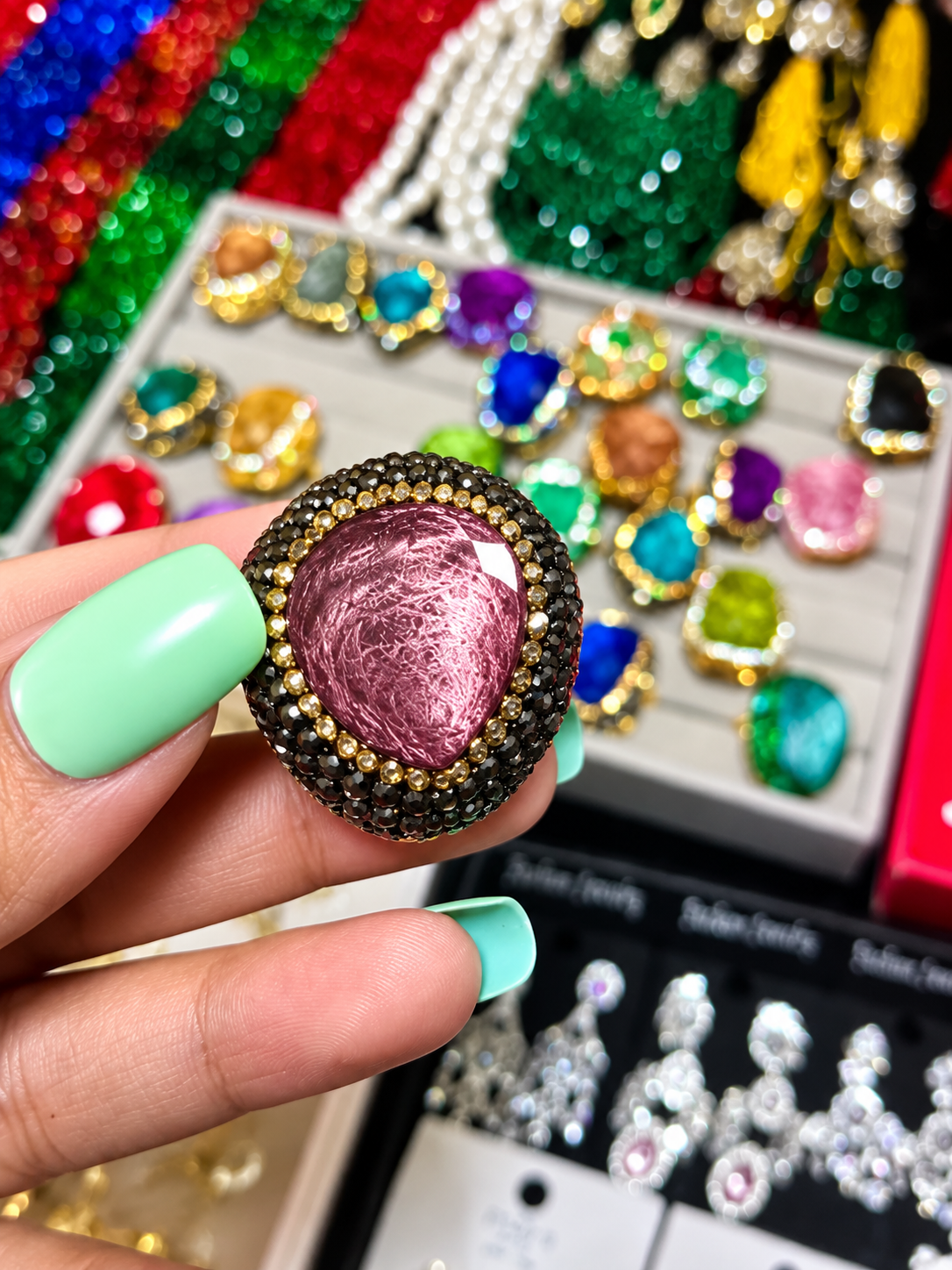 A hand with mint green nail polish holds a round fashion jewelry piece featuring a textured pink center surrounded by a dark brown beaded border, with a colorful display of other jewelry pieces in the blurred background.